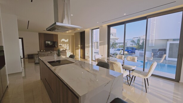 A modern kitchen with a large island, a dining area with chairs, and a view of a swimming pool and buildings outside. - 146 5 AN - Stay Oasis Luxury Villa (Ayia Napa)