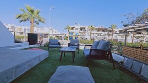An outdoor seating area with a table and chairs, a small patch of artificial grass, and a view of surrounding buildings.