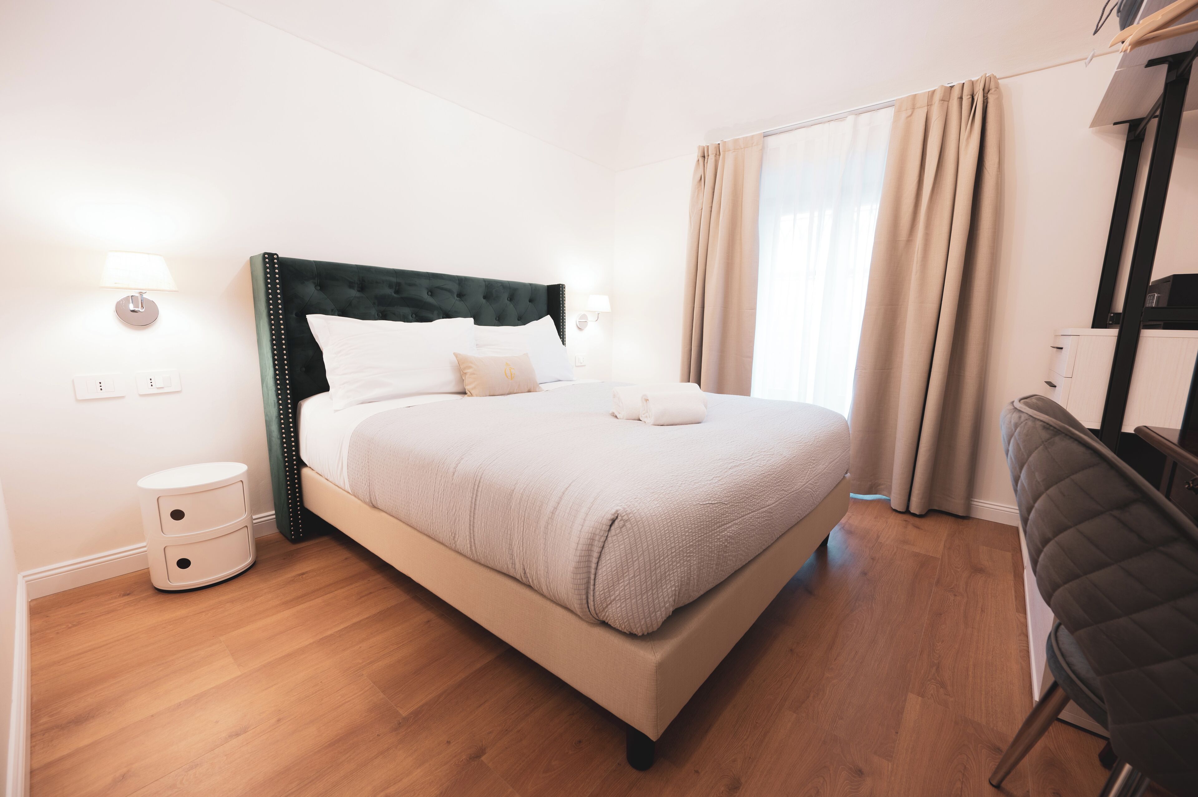 Una cama bien hecha con cabecera tapizada en verde, una mesita de noche con cajones y una silla a la derecha.