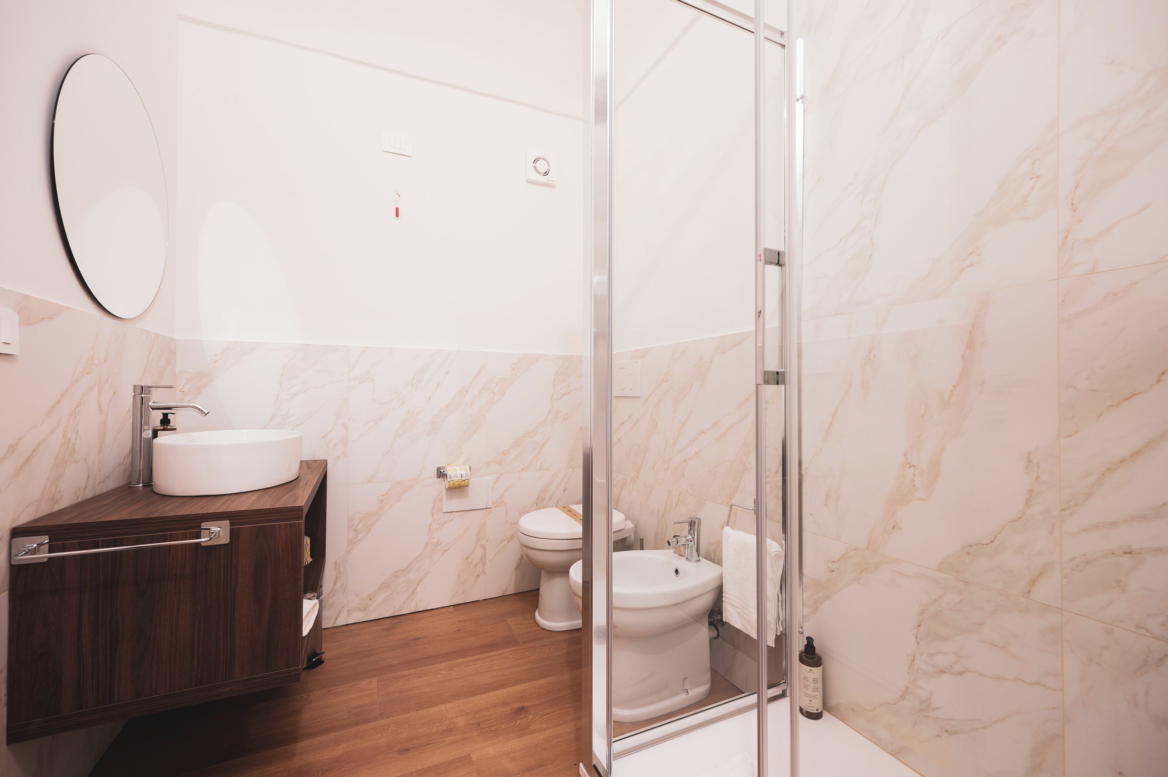 A modern bathroom with a marble wall, a wooden vanity, a glass shower enclosure, and a toilet.