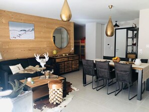 Un salon moderne avec un mur en bois, un miroir rond, un coin repas avec une table et des chaises, et une cuisine en arrière-plan.