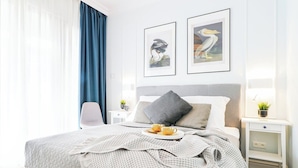 A neatly arranged bedroom with a bed, bedside tables, a nightstand, a window with curtains, and two framed pictures on the wall.