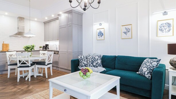 A modern living room with a teal sofa, white coffee table, and a dining area with white chairs and a kitchen in the background.