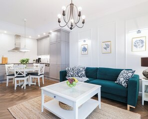 Una sala de estar moderna con un sofá color esmeralda, una mesa de centro blanca y un comedor con sillas blancas, con una cocina al fondo.