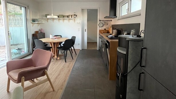 A modern kitchen with a dark countertop, a dining area with a wooden table and chairs, and a view through a glass door.