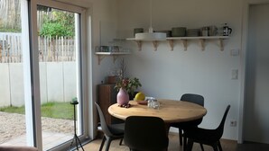 A dining area with a round table, chairs, a vase with flowers, and shelves above holding plates and cups.