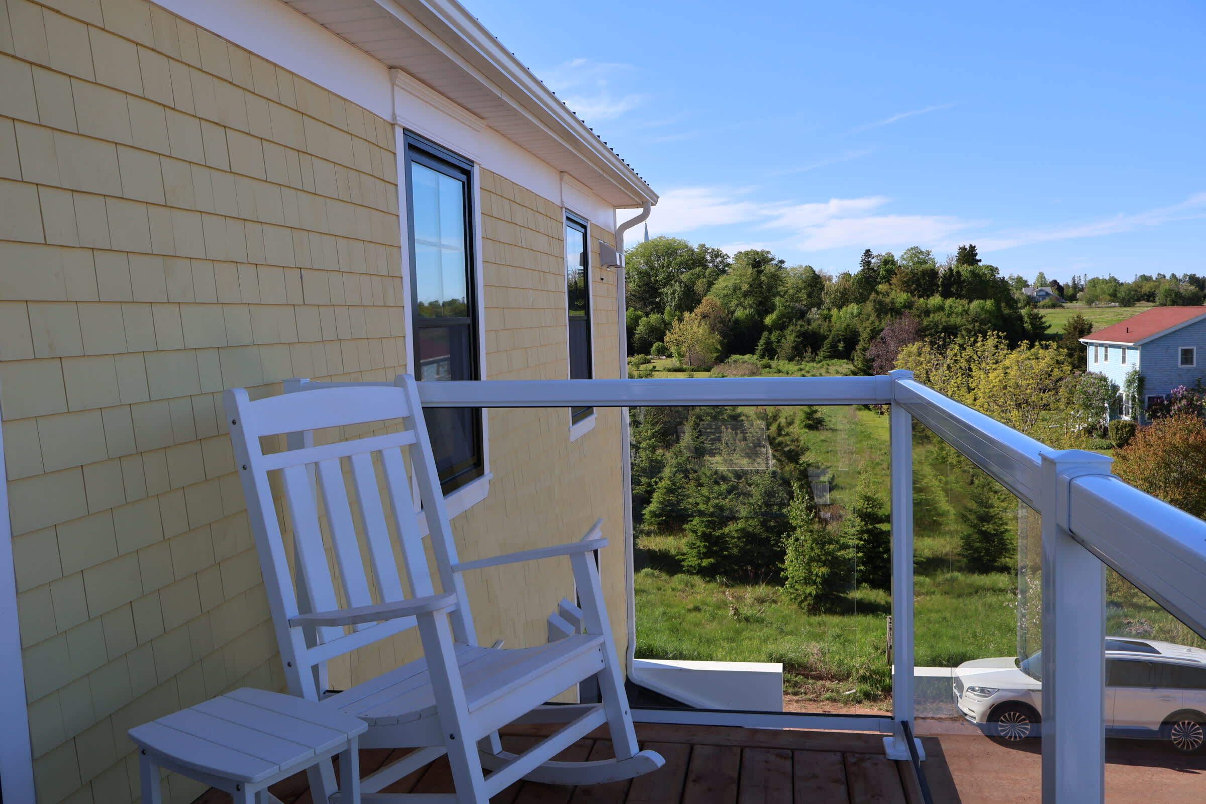 Une terrasse en bois avec un fauteuil à bascule et une balustrade blanche, d’où l’on peut admirer un paysage verdoyant et une maison.