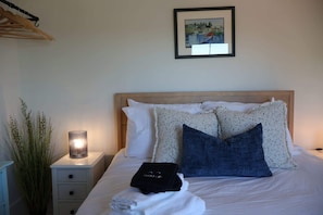 Ein ordentlich bezogenes Bett mit Kopfteil, eine Nachttischlampe, ein gerahmtes Bild an der Wand und ein kleiner Tisch mit Pflanze und Tasche.