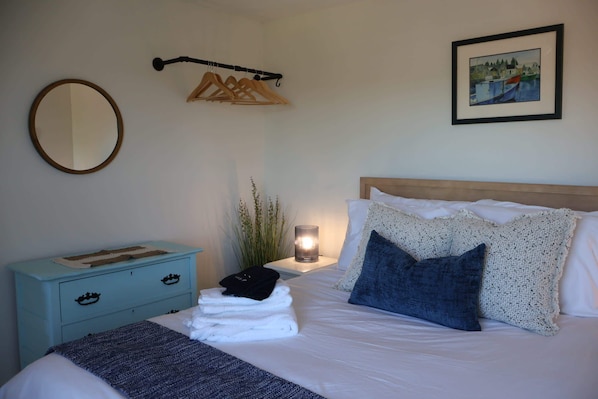 Ein ordentlich gemachtes Bett mit weißen Leinen und einem blauen Kissen, ein kleiner Nachttisch mit Lampe, ein wandmontierter Spiegel und ein gerahmtes Bild über dem Bett.