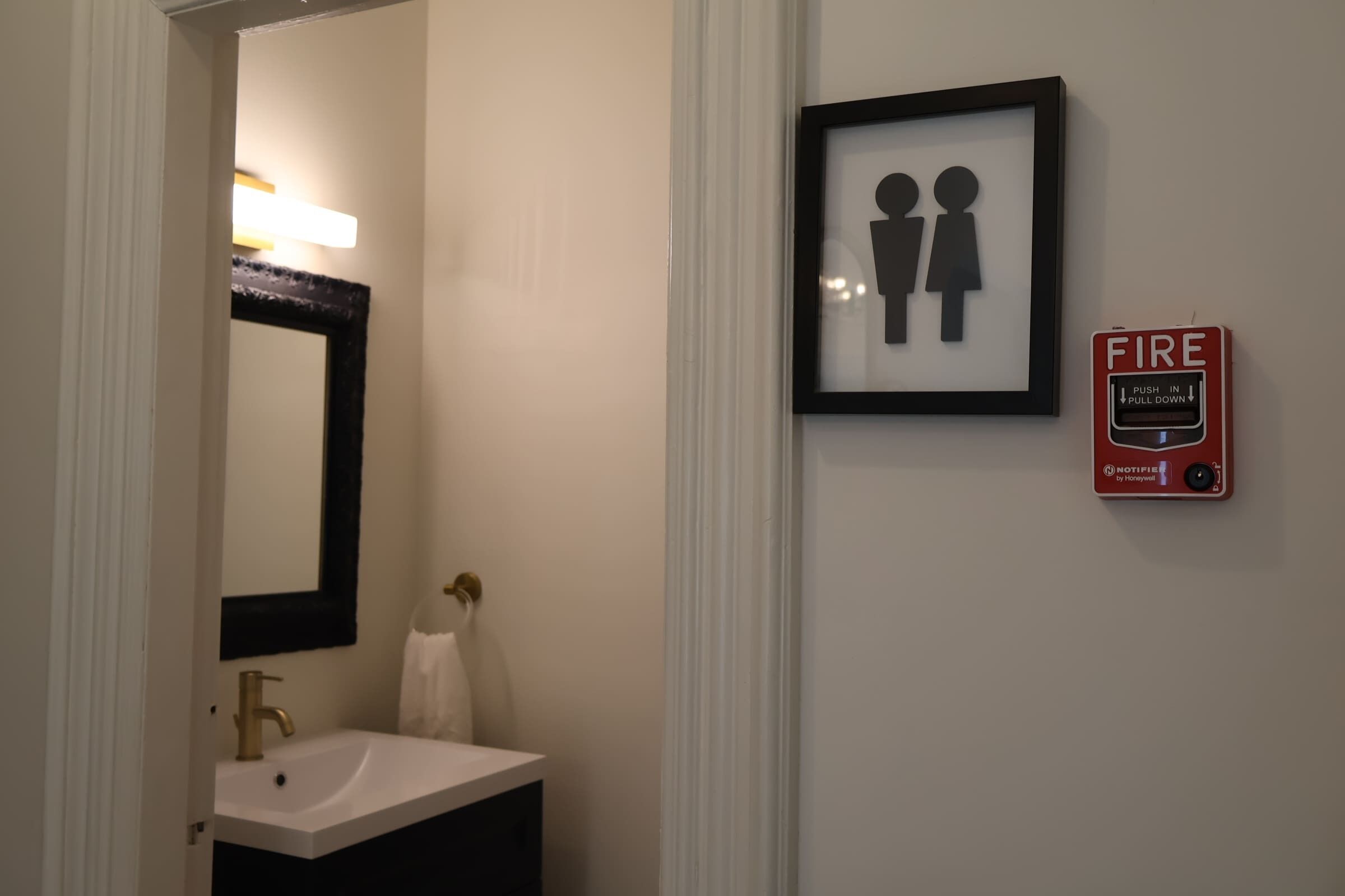 Une salle de bain avec un lavabo, un miroir, un tableau encadré représentant deux personnes et un détecteur d’incendie.