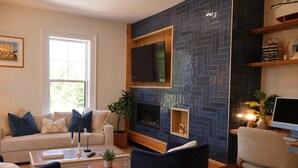 A living room with a sofa, coffee table, and TV. There's a fireplace with a TV above it, and a bookshelf with decorative items.