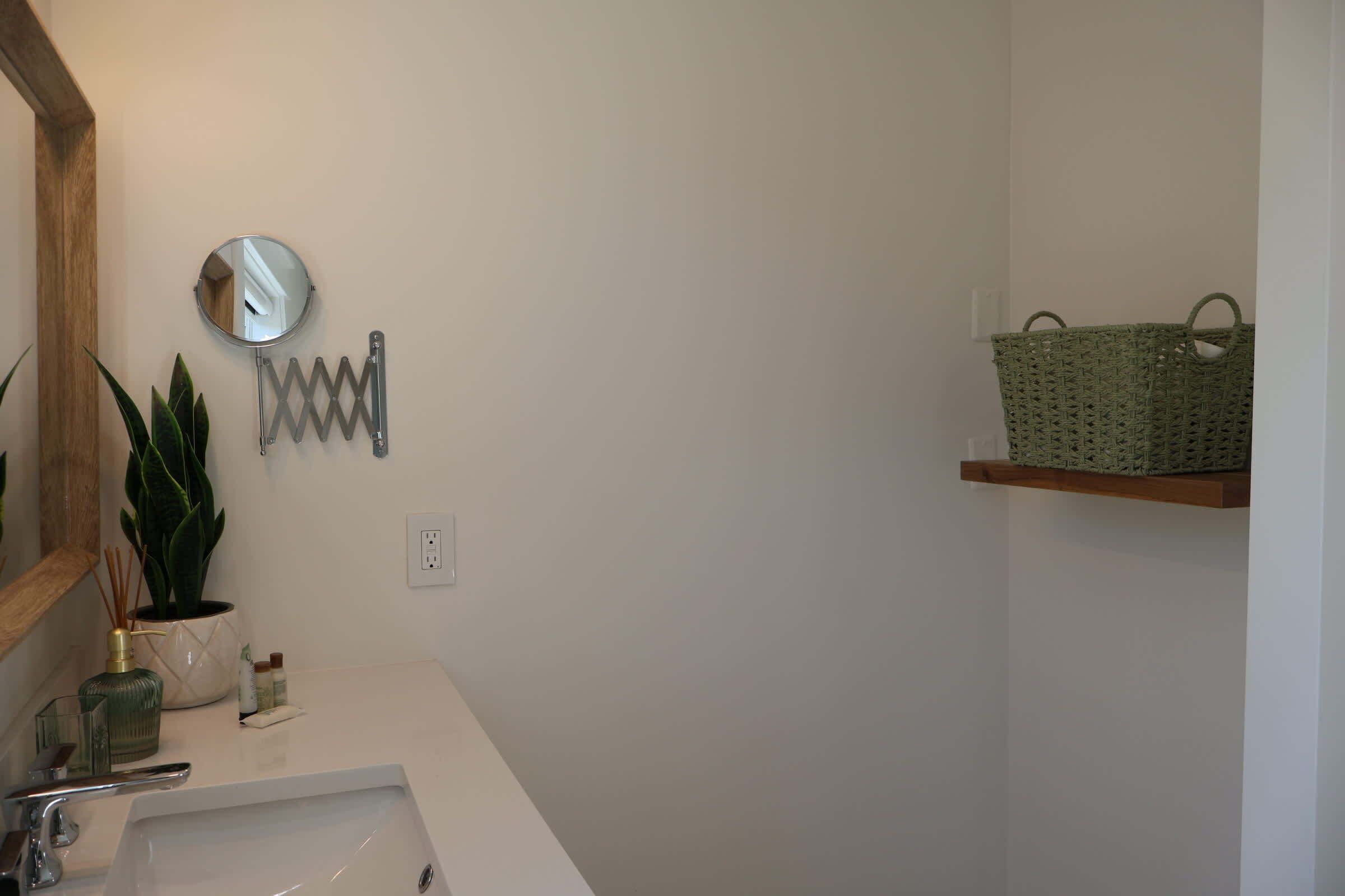 A bathroom with a white sink, a mirror, a green plant, a hanging towel rack, and a woven basket on a shelf.