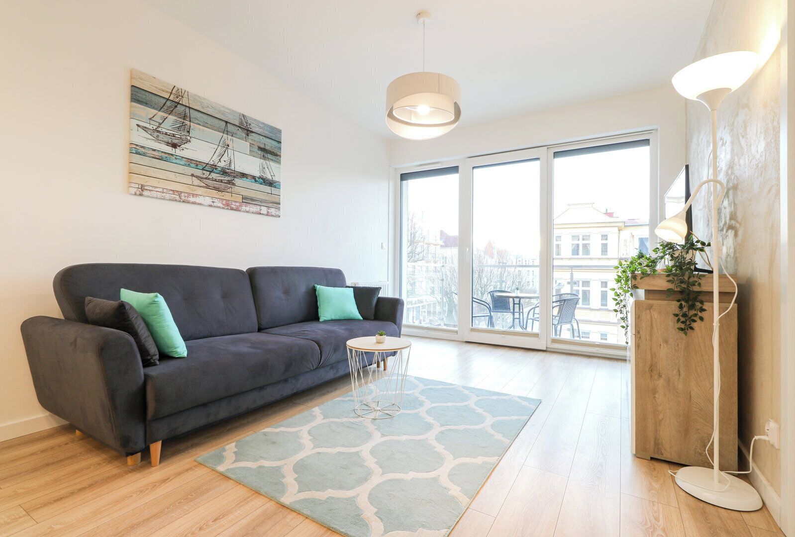 A modern living room with a dark grey sofa, a small round table, and a large window offering a city view.