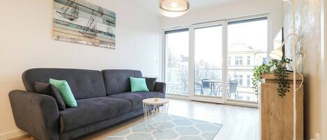 A modern living room with a dark grey sofa, a small round table, and a large window offering a city view.
