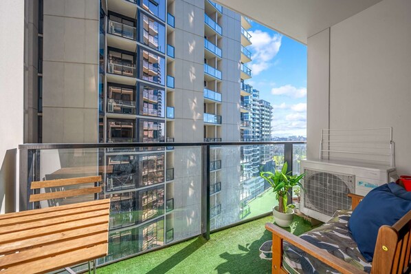 A balcony with a view of a high-rise building, a wooden bench, a small table with a plant, and a sofa with a cushion.