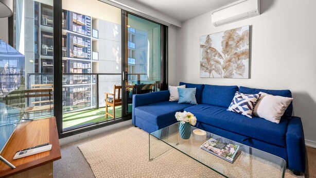 A modern living room with a blue sofa, a glass coffee table, and a view of a building through large windows.