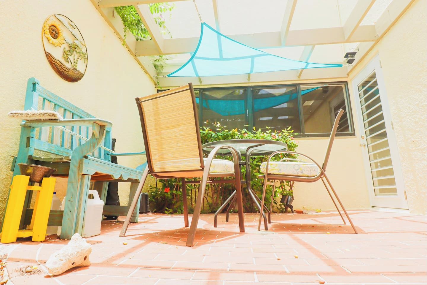 A patio with a table and chairs, a blue chair, a yellow chair, a wall-mounted decorative plate, and a window with a blue awning.