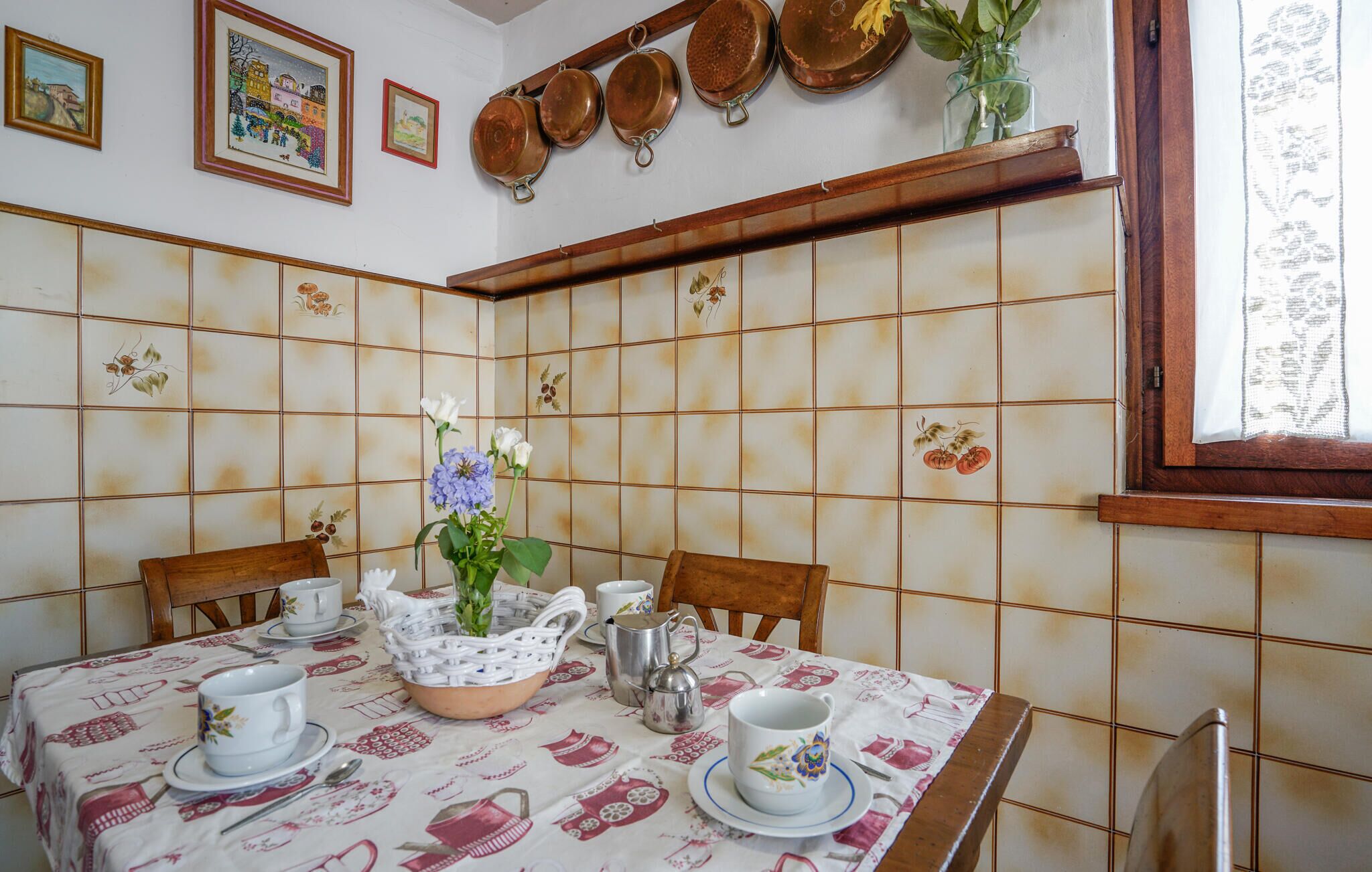 A dining area with a tiled wall, a table set with white cups and saucers, a metal pitcher, and a woven basket with flowers.