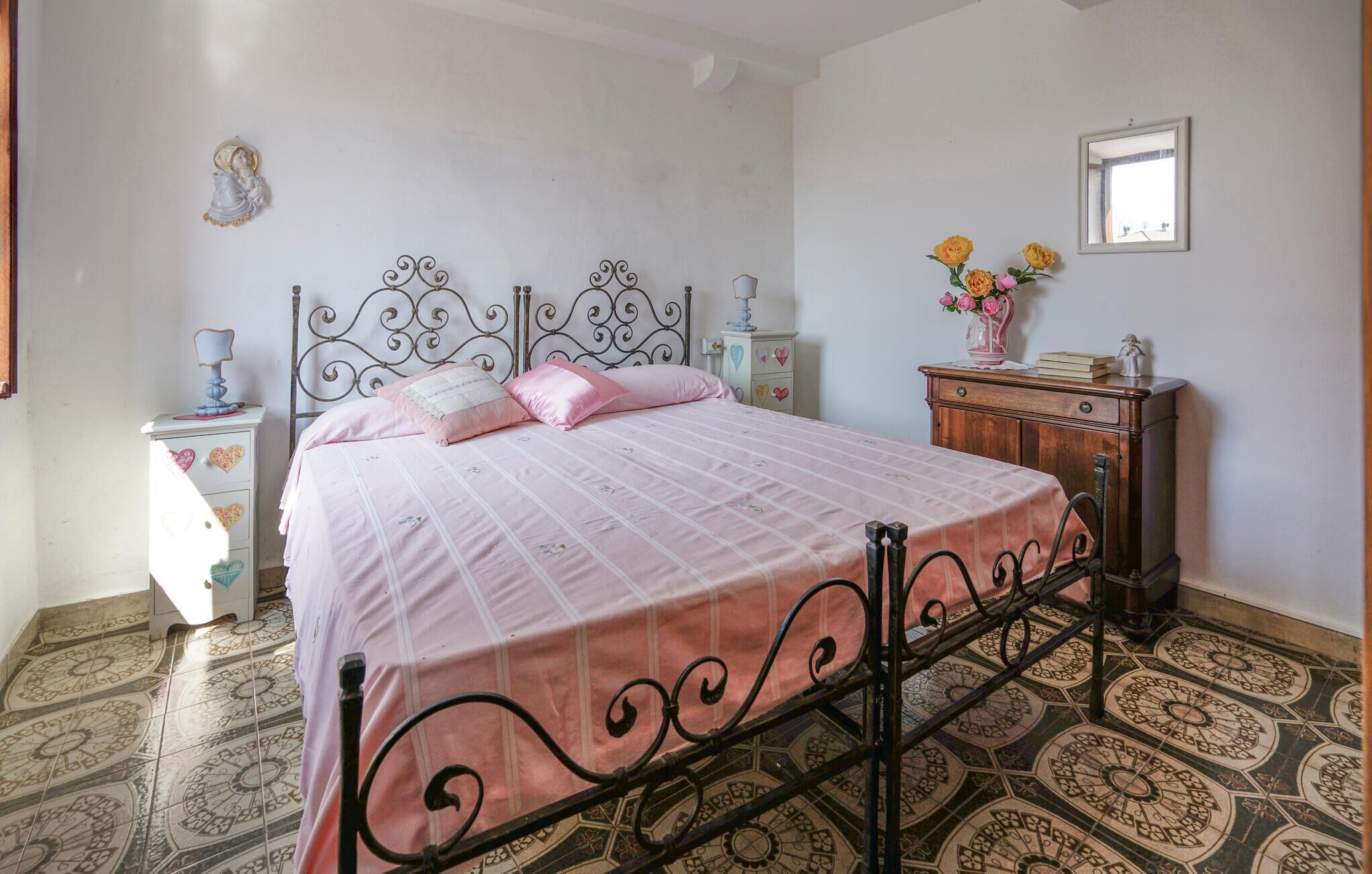 A bedroom with a bed, a nightstand, a dresser, a mirror, and a vase with flowers.