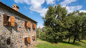 A stone building with wooden shutters, a stone pathway, and a large tree in the background.