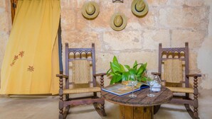 Una habitación rústica con una mesa de madera, dos sillas, un cortinón amarillo y tres sombreros de paja en la pared.