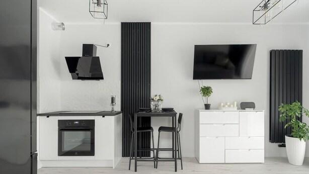 A modern kitchen with a built-in oven, a dining area with two chairs, a white cabinet, a flat-screen TV mounted on the wall, and a potted plant.