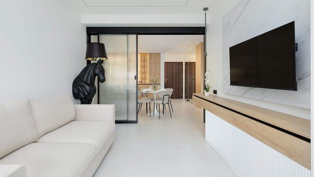 A modern living room with a white sofa, a black lamp with a horse design, a dining area with a white table and chairs, and a mounted TV on the wall.