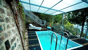 A swimming pool with a glass enclosure, a stone wall, and a staircase leading to a lounge area.