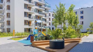 Un immeuble d’appartements moderne doté d’un espace de jeux pour enfants, comprenant des toboggans et une structure d’escalade, le tout entouré de verdure et agrémenté d’une fontaine.