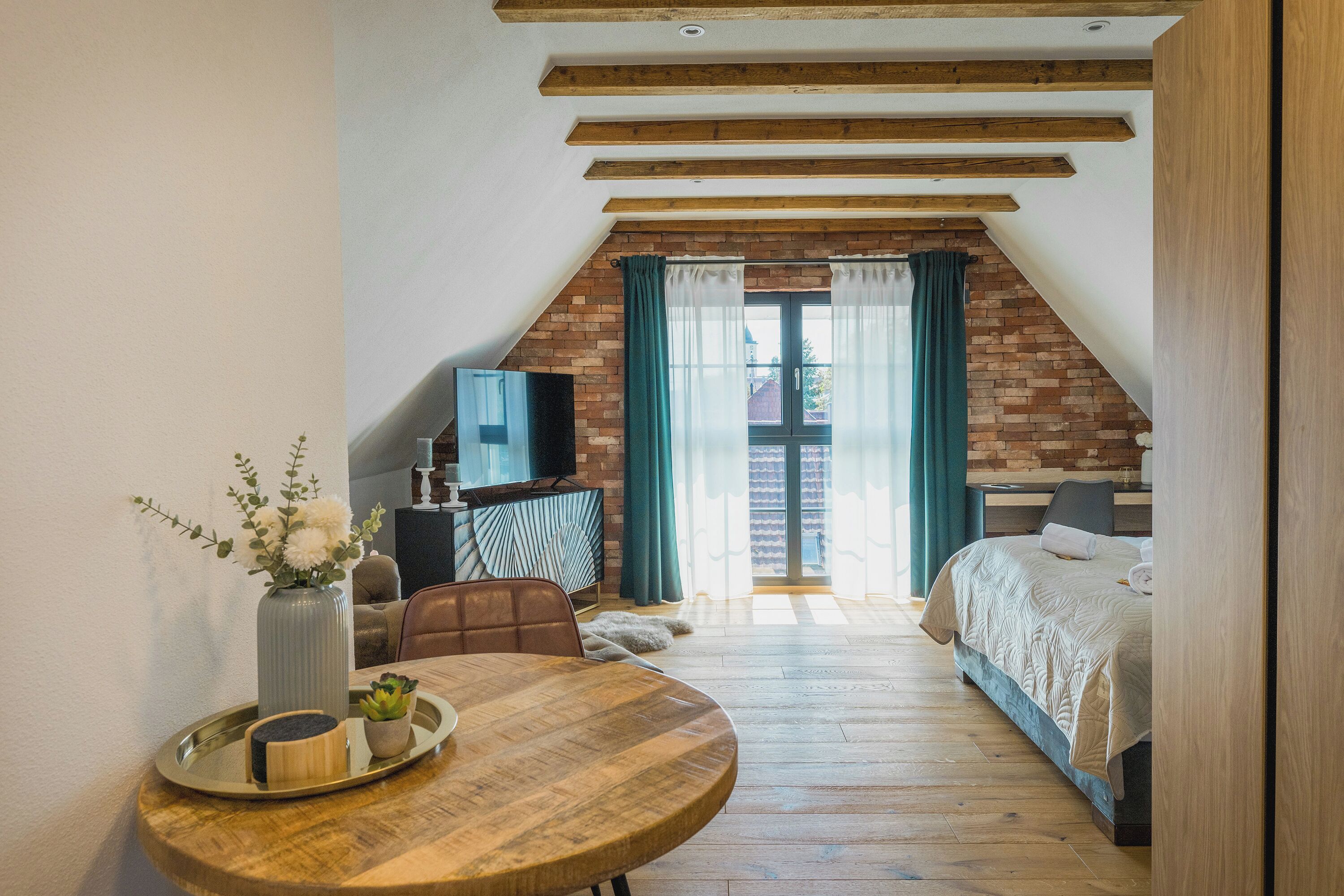 A cozy attic bedroom with a wooden ceiling, a brick wall, a bed, a TV, and a dining area with a table and chairs.