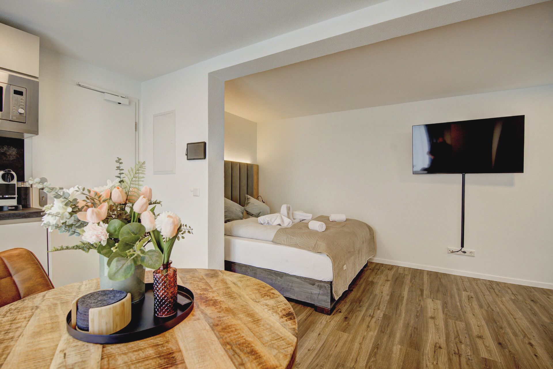 Una habitación de hotel moderna con suelo de madera, una cama con manta color crema, un televisor montado en la pared y una zona de comedor con mesa de madera y un jarrón de flores.