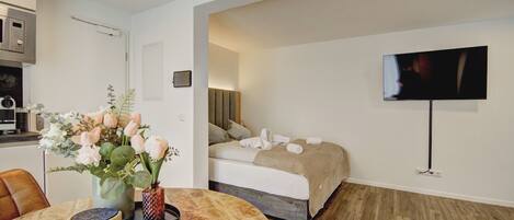 Una habitación de hotel moderna con suelo de madera, una cama con manta color crema, un televisor montado en la pared y una zona de comedor con mesa de madera y un jarrón de flores.