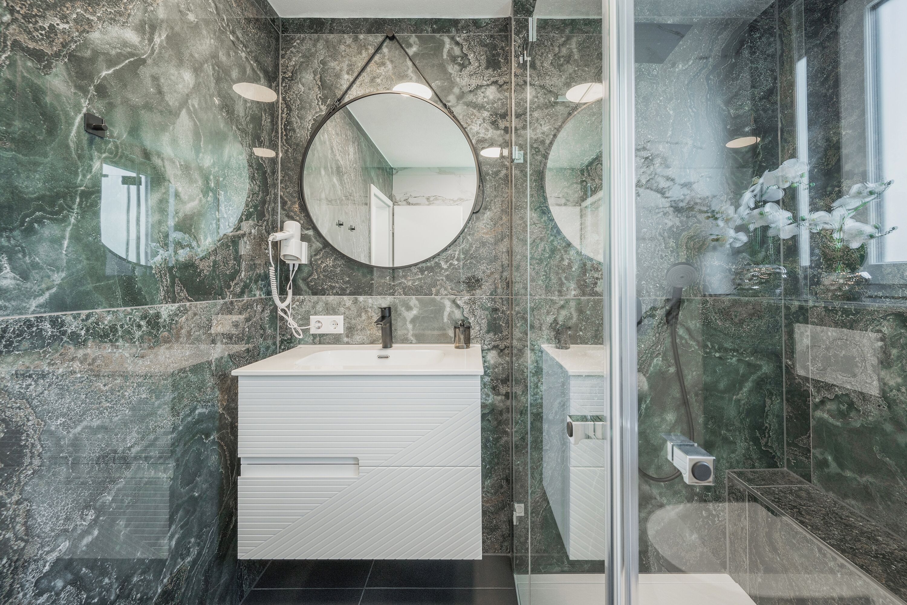 A modern bathroom with a marble wall, a round mirror, a white vanity, and a glass shower enclosure.
