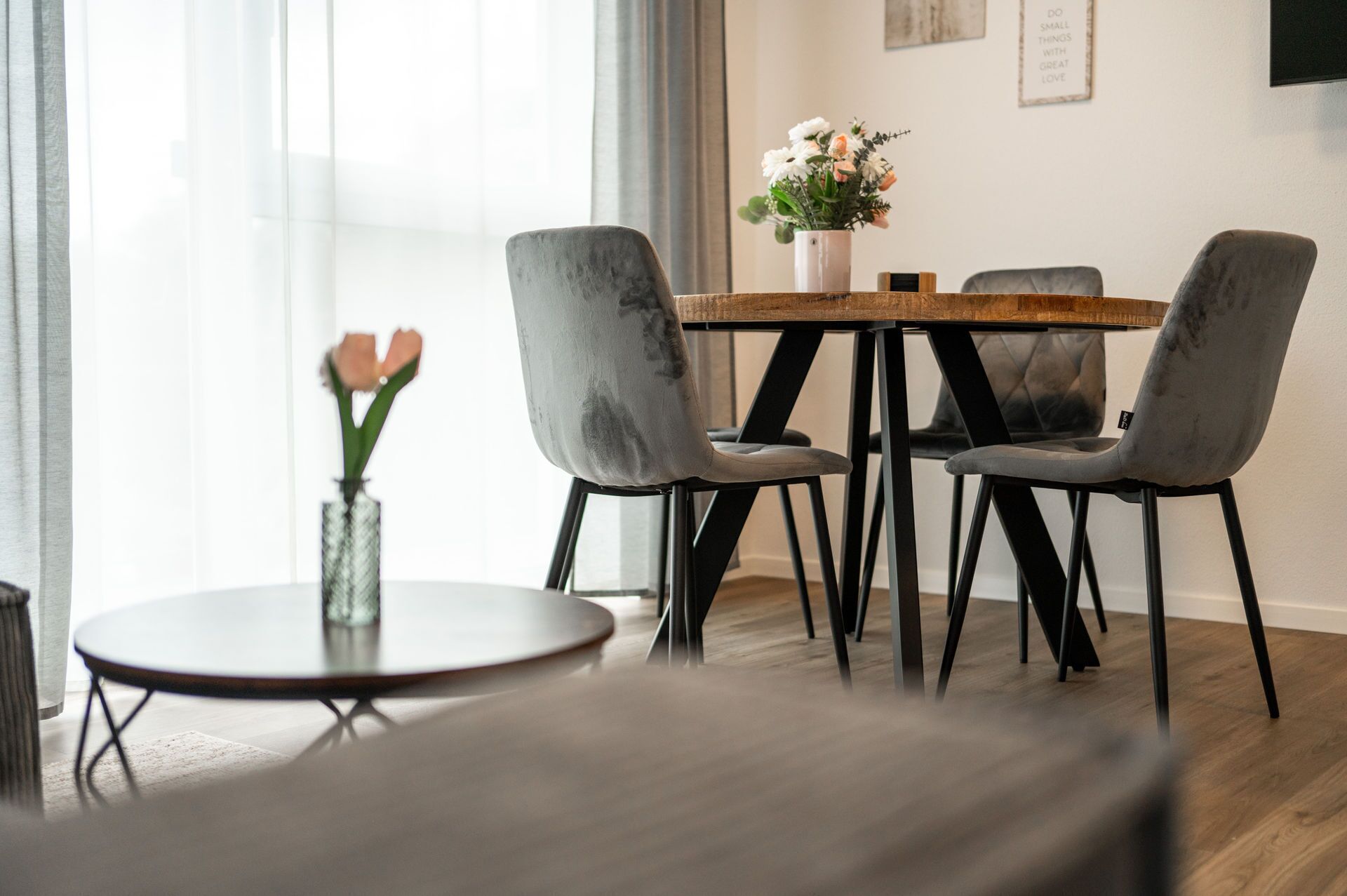 A modern dining area with a round wooden table and four grey upholstered chairs. A vase with flowers is on the table, and a single rose is in a vase on the floor.