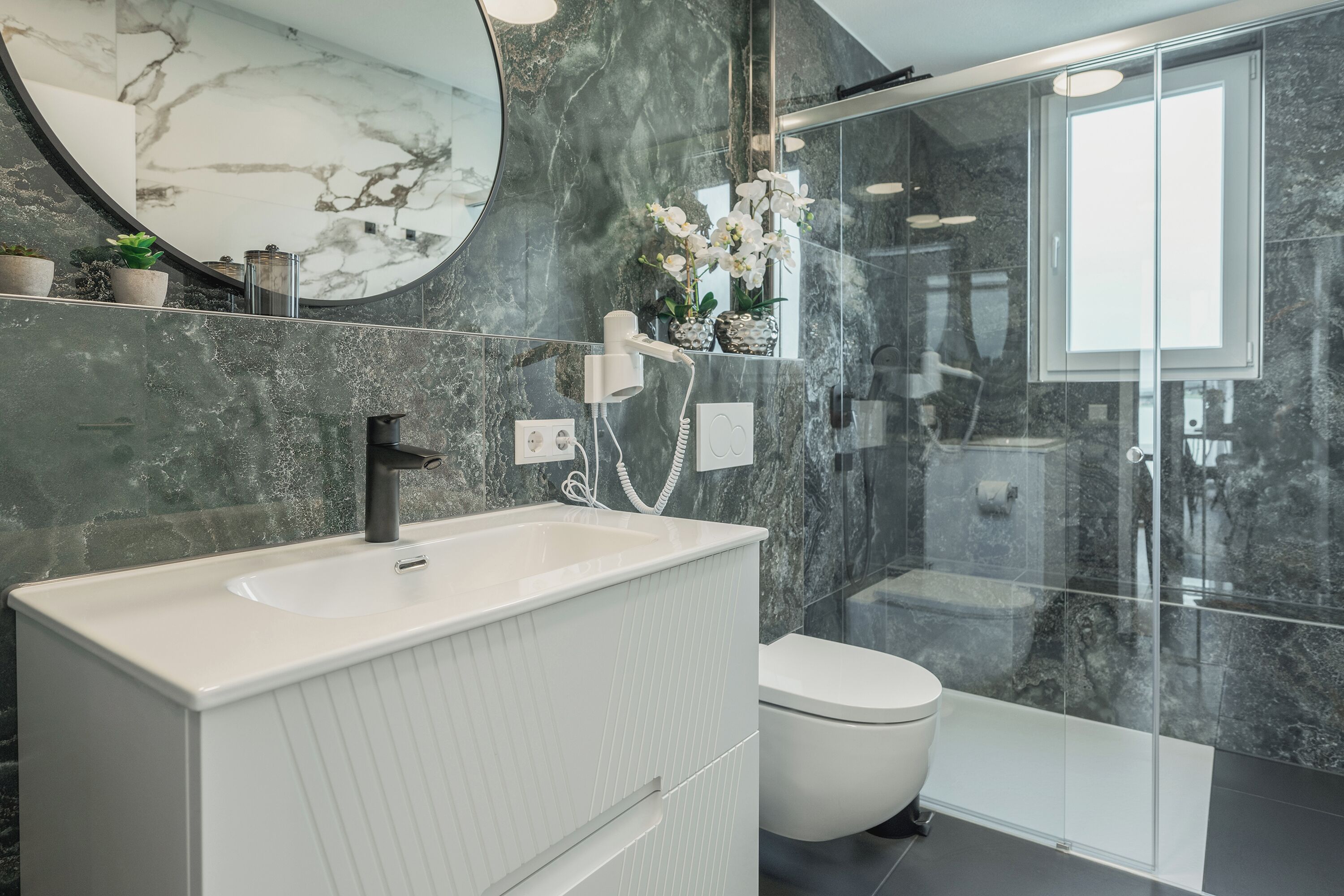 A modern bathroom with a white vanity, a round mirror, a toilet, and a glass-enclosed shower area.
