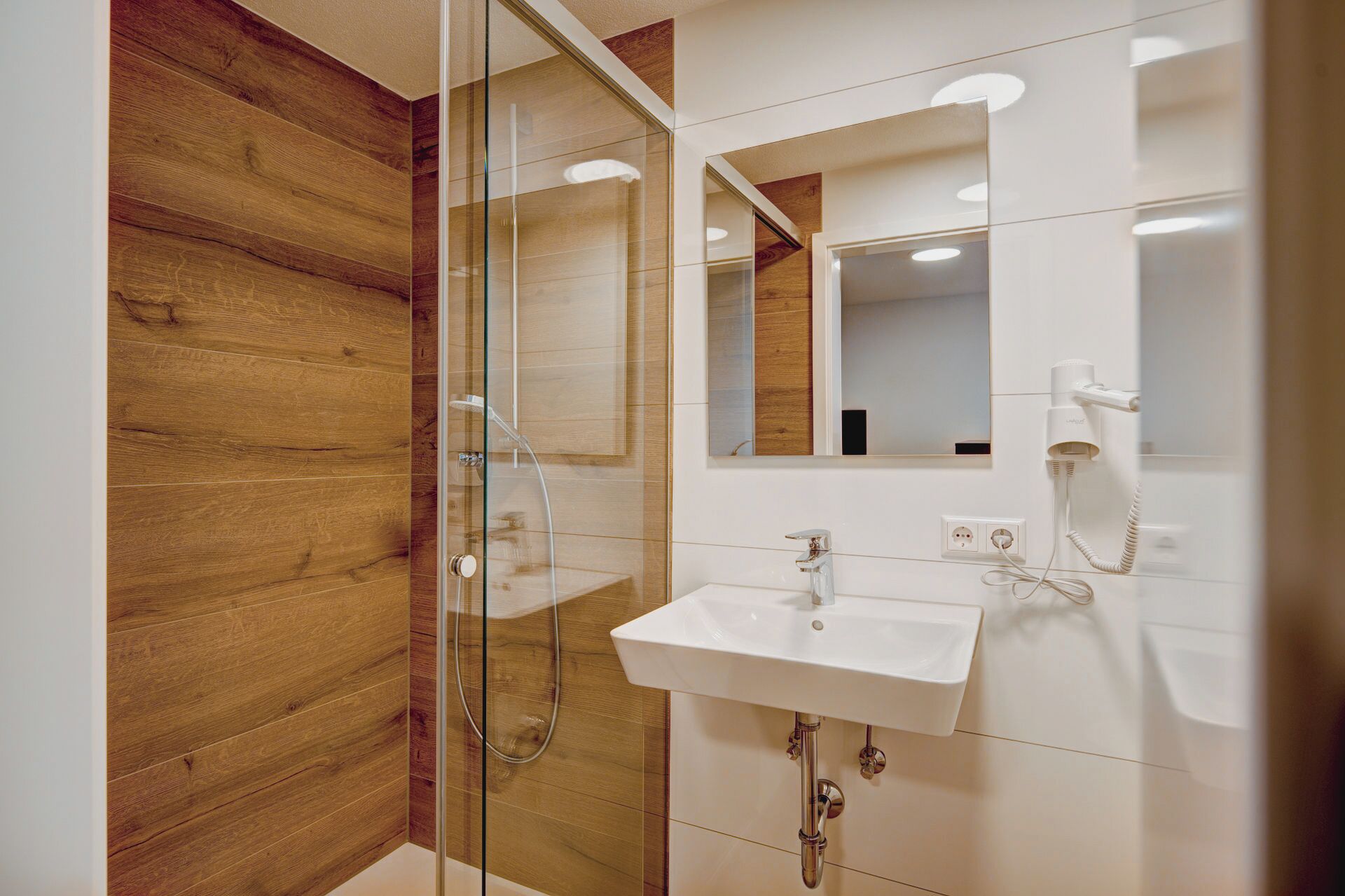 A modern bathroom with a wooden accent wall, a glass shower enclosure, a white sink, and a hairdryer.