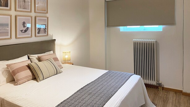 Une chambre bien rangée, avec un lit, des tableaux encadrés accrochés au mur, un appareil de climatisation et une fenêtre équipée de stores.