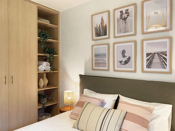 Ein Schlafzimmer mit Bett, Kissen, Nachttisch, Lampe, Kleiderschrank und gerahmten Bildern an der Wand.
