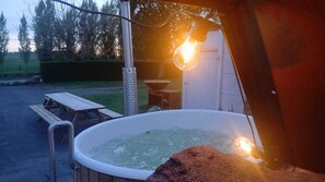 Un jacuzzi con un banco de madera y una mesa cerca, rodeado por un jardín y una caseta.