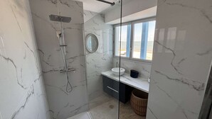 Un baño moderno con ducha de vidrio, lavamanos con espejo y vista a la playa a través de una ventana.