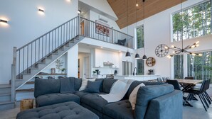 Un amplio salón con un sofá seccional gris grande, una escalera de madera y un comedor con mesa y sillas de madera.