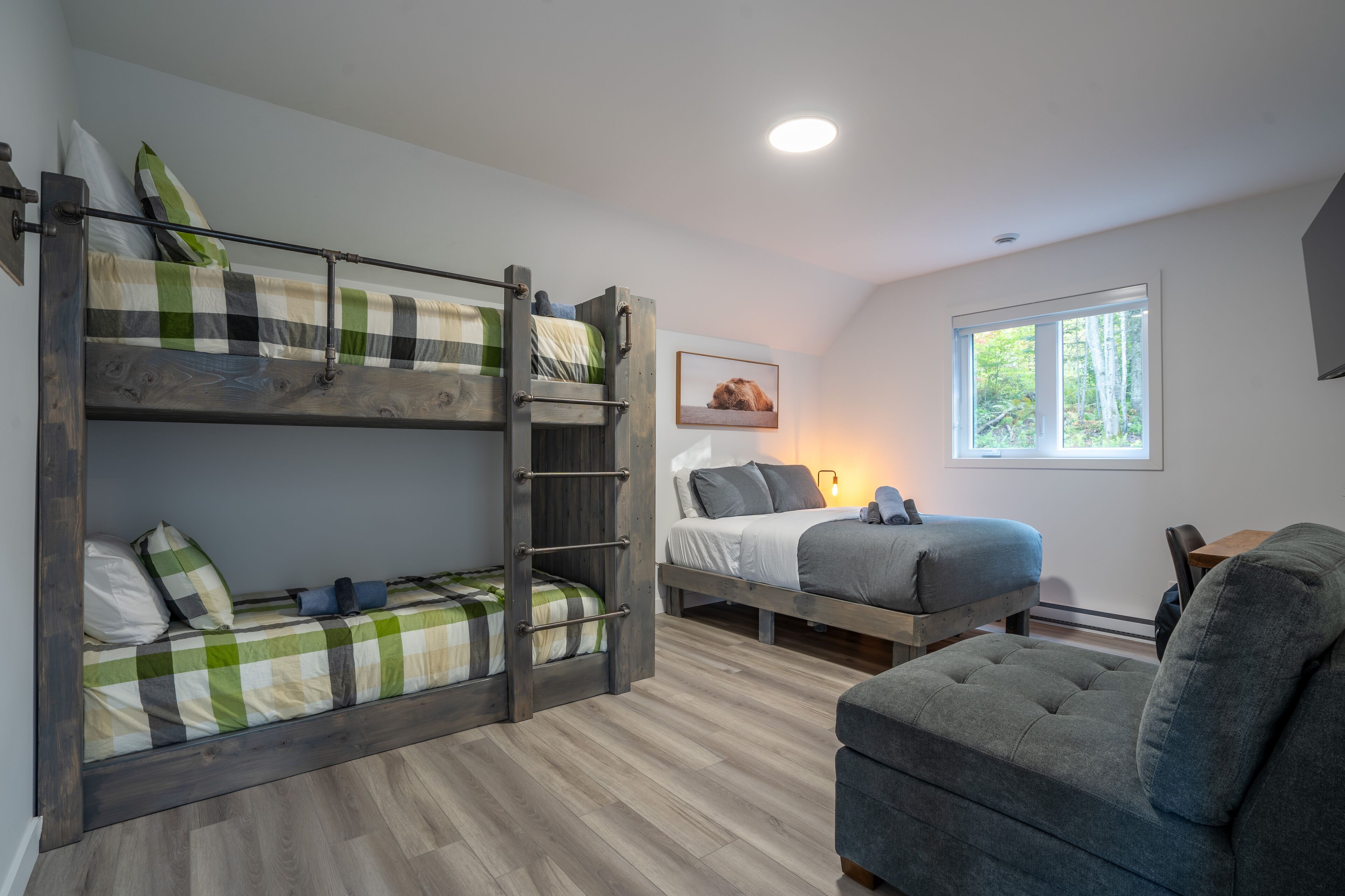 Un lit superposé avec une échelle, un lit simple, un canapé gris et une fenêtre donnant sur des arbres.