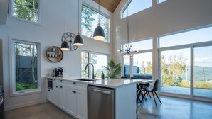 Una cocina moderna con armarios blancos, lavavajillas de acero inoxidable y una isla grande. Varias ventanas ofrecen luz natural y vistas al exterior.