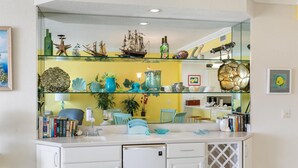 Une cuisine avec des meubles blancs, une étagère en verre ornée d’objets décoratifs et un mur jaune.