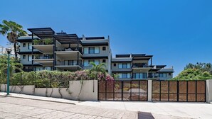 Un moderno edificio residencial de varios pisos con balcones, entrada vigilada y abundante vegetación.