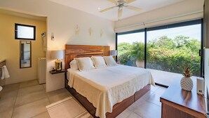 Un dormitorio amplio con una cama grande, un ventilador de techo, vista a la vegetación a través de una puerta corredera y un pequeño escritorio con una decoración de piña.