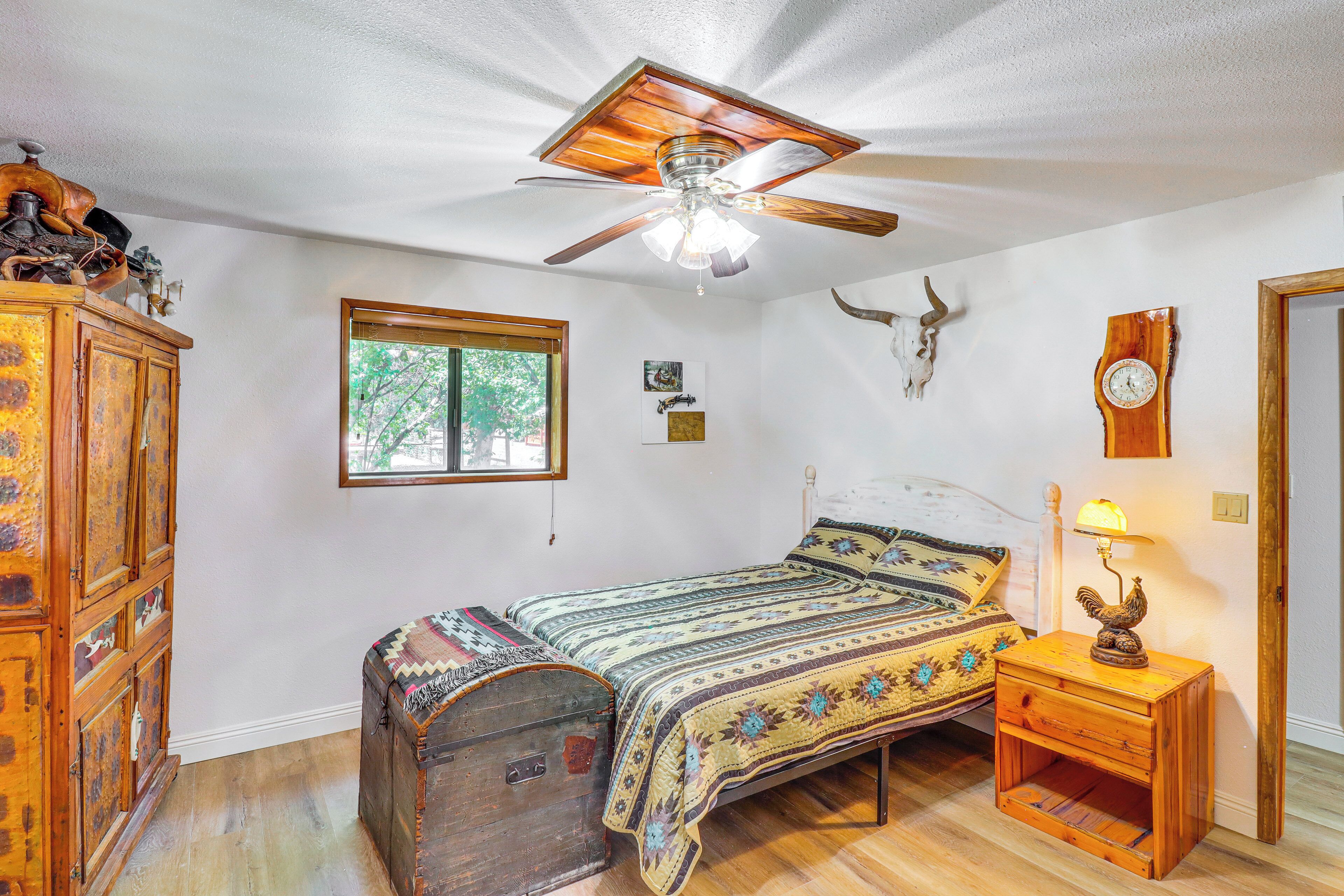Ein Schlafzimmer mit einem hölzernen Bett, einem Deckenventilator, einer rustikalen Kommode, einer an der Wand befestigten Uhr und einem Fenster mit Blick auf Bäume.