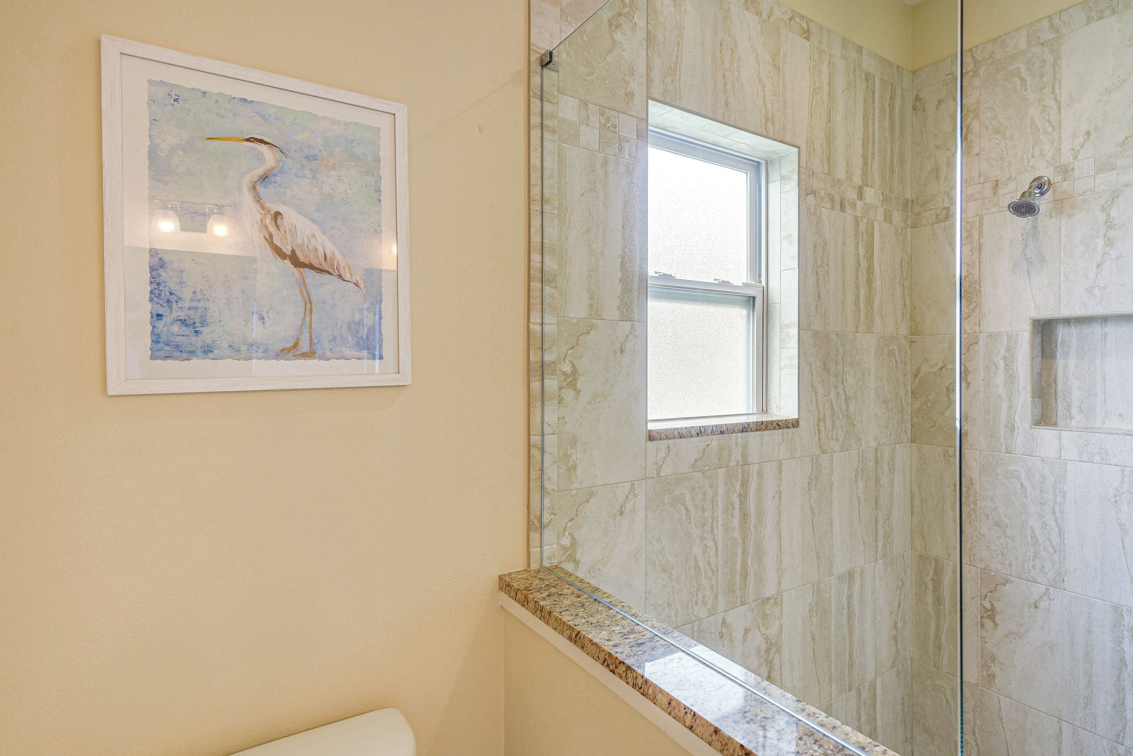 A bathroom with a marble wall, a framed painting of a bird, and a window.