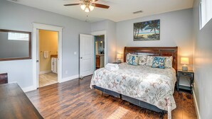 A bedroom with a wooden floor, a ceiling fan, a bed with a floral bedspread, a nightstand with a lamp, and a painting of a beach scene. - Close to Beaches: Palm Coast Home w/ Deck! (Palm Coast)