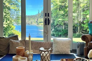 A room with a large window offering a view of a lake and forest. There is a sofa, a table with candles, and a chair. - Bayfront PNW Lodge: Dock, Pickleball & Guest House (Bainbridge Island)